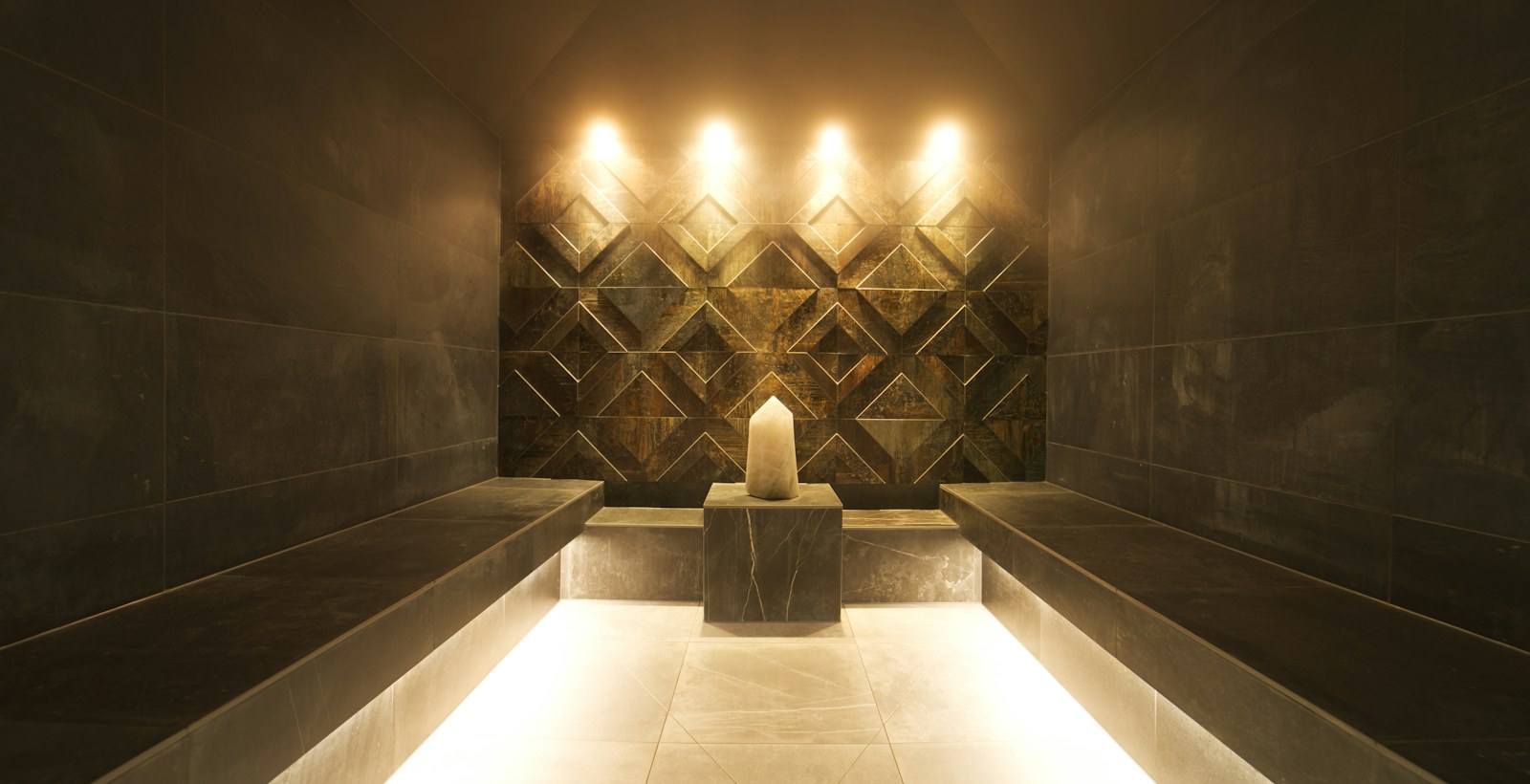 Lamplit stone steam room with tiled benches and a low central fountain